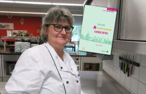 Eine Frau in Kochkleidung steht in einer Großküche und lächelt in die Kamera. Neben ihr ist ein Cover mit dem Titel „Vegane & Vegetarische Gerichte“ eingeblendet.