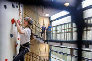 Eine Person mit Helm und Kletterausrüstung steigt eine Indoor-Kletterwand hinauf.