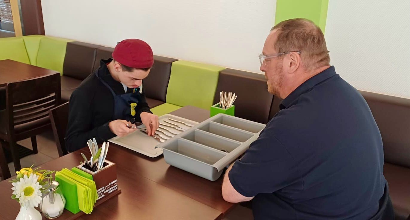 Ein junger und ein älterer Mann sitzen gemeinsam an einem Tisch. Der junge Mann legt Messer gerade vor sich auf ein Platzset.