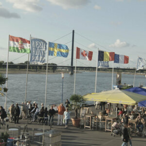 Das Bild zeigt die Düsseldorfer Rheinuferpromenade mit vielen Menschen, Sonnenschirmen und Fahnen. Im Hintergrund ist die Oberkasseler Brücke zu sehen.