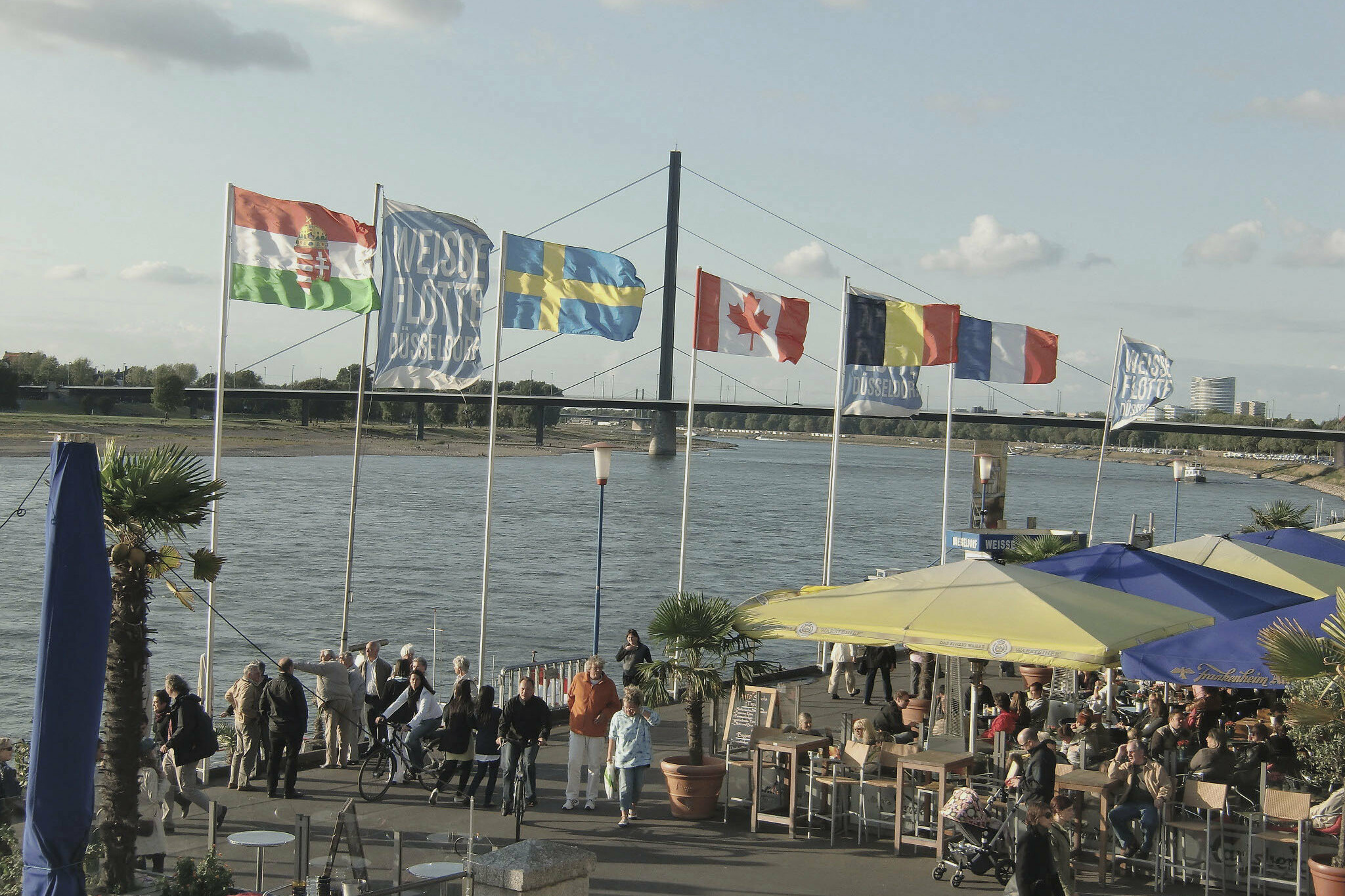 Das Bild zeigt die Düsseldorfer Rheinuferpromenade mit vielen Menschen, Sonnenschirmen und Fahnen. Im Hintergrund ist die Oberkasseler Brücke zu sehen.