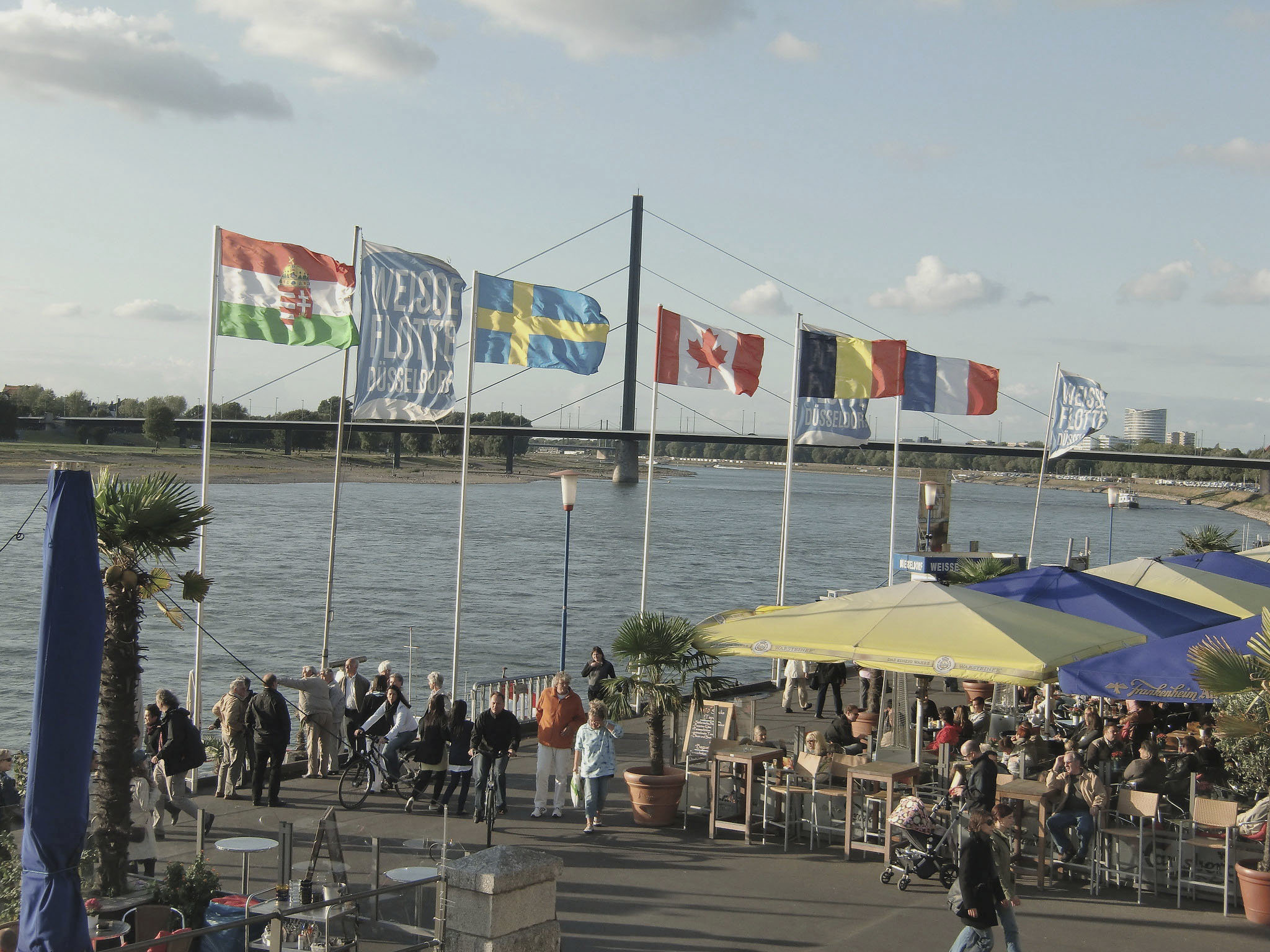 Das Bild zeigt die Düsseldorfer Rheinuferpromenade mit vielen Menschen, Sonnenschirmen und Fahnen. Im Hintergrund ist die Oberkasseler Brücke zu sehen.