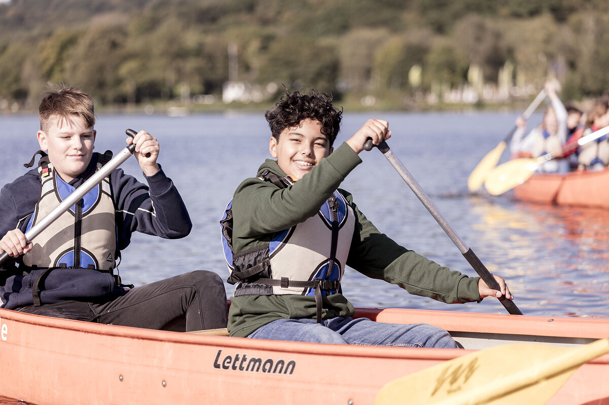 Das Bild zeigt zwei Jungen, die auf einem See Kanu fahren. Im Hintergrund ist ein weiteres Kanu zu sehen.