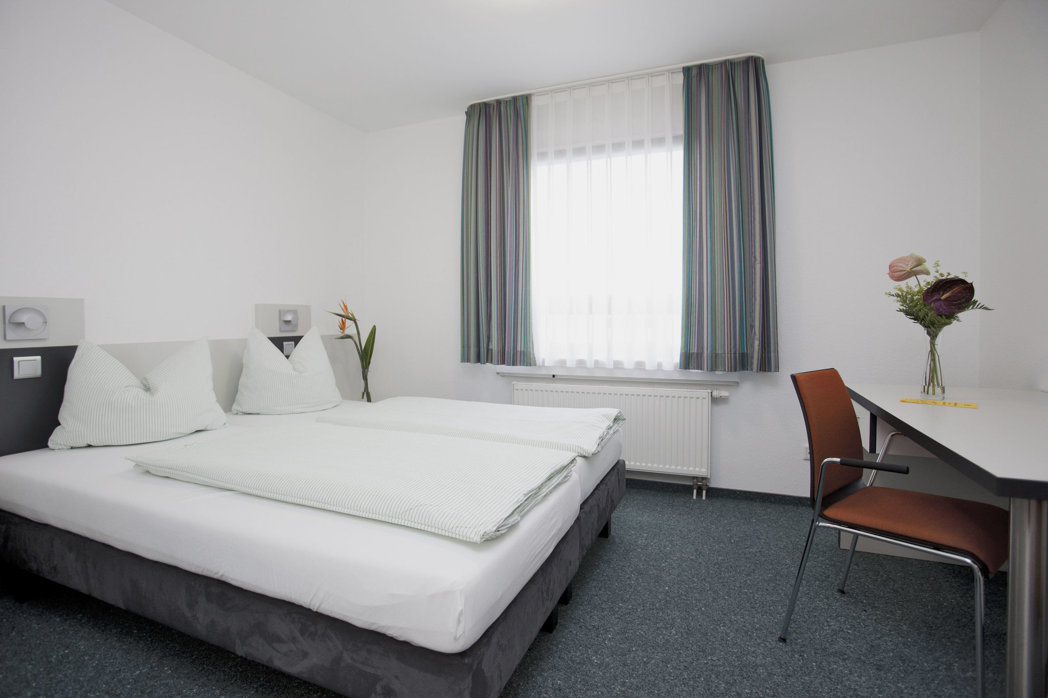 Ein Doppelbett vor einem Fenster mit Gardinen, daneben ein Stuhl