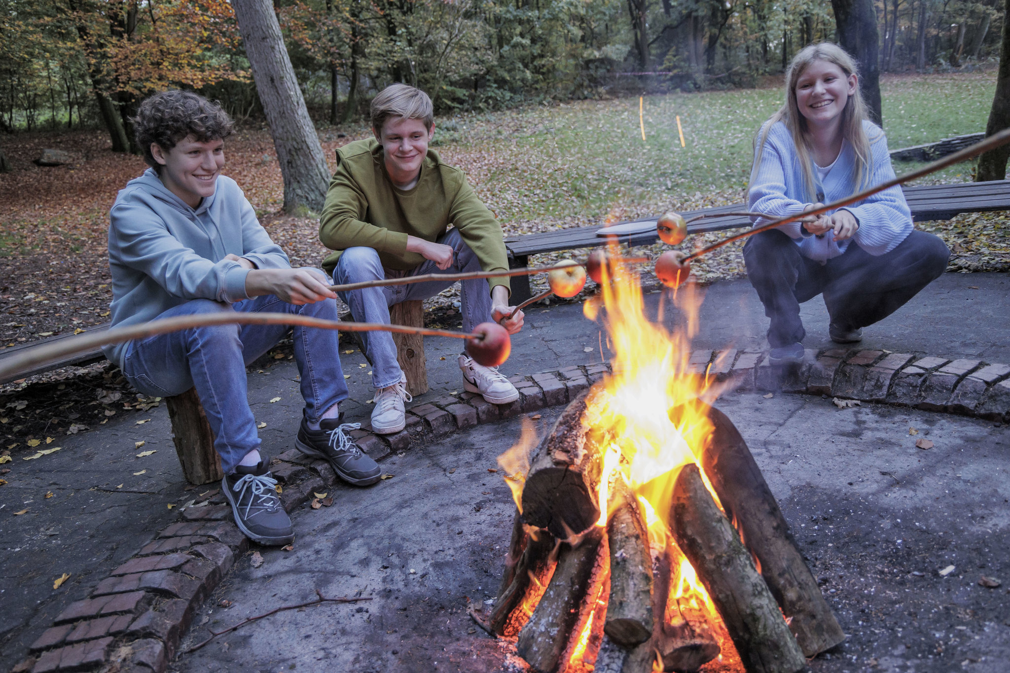 Drei Jugendliche - zwei Jungs, ein Mädchen - backen in der Dämmerung Äpfel am Lagerfeuerpfel