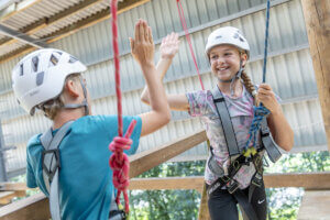 Zwei Kinder mit Klettergurten und Helmen geben sich ein High-Five auf einem Hochseilgarten-Parcours.