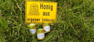 Drei Honiggläser stehen vor einem gelben Schild mit der Aufschrift „Honig aus eigener Imkerei“ auf einer Wiese.