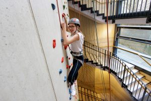 Person mit Helm und Klettergurt an einer Kletterwand. Im Hintergrund sind Treppen und Geländer zu sehen.