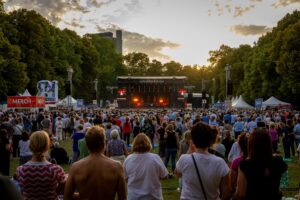 Viele Menschen stehen vor einer großen Konzertbühne im Park bei Dämmerung.