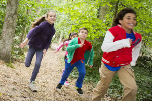 Vier Kinder rennen einen Waldweg entlang, umgeben von hohen Bäumen