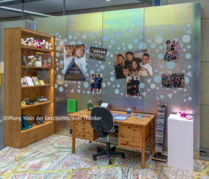 Ein Kinderzimmer, das an die 90er/00er Jahre erinnert. An der Wand über einem Schreibtisch hängen Poster der angesagten Boy- und Girlbands und die Diddl-Maus.