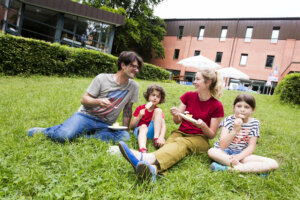 Eine vierköpfige Familie sitzt im Gras vor einem roten Haus. Die Eltern essen Kuchen, die Kinder ein Eis.