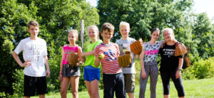Eine Gruppe von Kindern steht auf einer Wiese zusammen. Die Kinder tragen Sportkleidung, einige haben Baseballhandschuhe an.