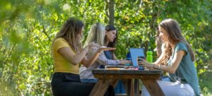 Mehrere Frauen sitzen im Wald am Picknicktisch und unterhalten sich. Auf dem Tisch stehen Laptops und Trinkflaschen.