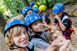 Eine Gruppe von Kindern sitzt nebeneinander im Wald. Sie tragen blaue Kletterhelme. Die beiden vorderen Jungen blicken lächelnd in die Kamera